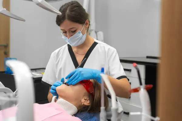 Female dentist in a uniform, medical mask examines the teeth of a patient in a dental clinic. Attractive patient lying on a dental chair receives dental treatment from a dentist.