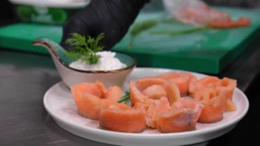 Salamura edilmiş somon parçalarını soslu ve dereotlu beyaz tabakta yakın plan çek. Güzel bir porsiyon salamura somon. Eldiven şefinin eli tabağı döndürür ve masaya koyar..