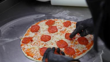 Profesyonel şef modern restoran mutfağında pizza hazırlıyor. Elleri siyah eldivenli. Pizzaya sosis salamı dilimleri seriyor.