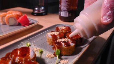 Yakın plan aşçı sıcak Banzai suşisine susam tohumu serperek somon, pirinç, krem peynir, susam tohumu, süslemeli tabakta teriyaki soslu panko, seçici odaklı. Yiyecek kavramı