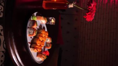 Asya restoranındaki güzel atmosferde, içinde çiçekler ve yanan mumlar bulunan yuvarlak tabakta pişmiş suşiler. dikey video