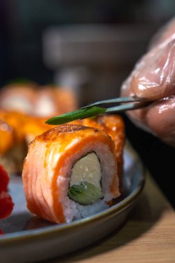 Eldivende cımbız kullanarak aşçının elinin yakın plan görüntüsü suşi rulolarına yeşil soğan, süzme peynir, balık ve salatalık koyuyor. Dikey fotoğraf