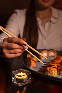 Genç bir kadın lüks bir restoranda taze suşi yer ve tadını çıkarır. Kız ellerinde çubuk tutuyor ve nefis bir suşi yiyor. Geleneksel Japon yemeği. Yemek kavramı. Dikey fotoğraf