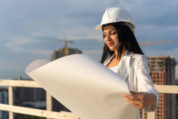 Kasketli genç ve güzel bir mimar güneşli bir günde inşaat alanındaki hangi adamın kağıdındaki çizimlere bakıyor. İnşaat müfettişi.