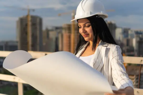 Kasketli genç ve güzel bir mimar güneşli bir günde inşaat alanındaki hangi adamın kağıdındaki çizimlere bakıyor. İnşaat müfettişi.