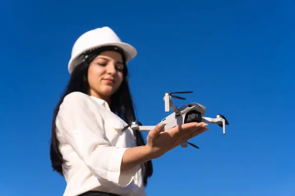 Kasklı genç bir kadın uzman mavi gökyüzü arka planına karşı elinde bir dron tutuyor. İnşaat ve endüstri kavramı