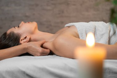 Masör, ön planda bulanık bir mum ve yeşil bir bitki olan kaplıcadaki kanepede uzanan güzel bir kadına derin bir kas masajı yapar.