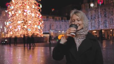 Mutlu gülümseyen bir kadının portresi, açık havada bir bardak sıcak içecekle Noel marketi, sokaklarda ve arka planda tezgahların yanında insanlar. Sokak Gecesi Şehir Işıkları. Dinlenme, rahatlama..