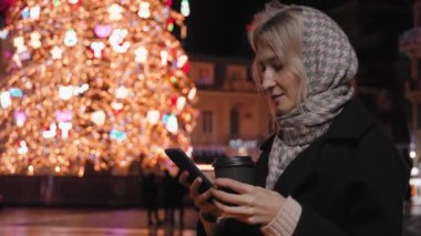 Genç ve çekici bir kadın akıllı telefon kullanıyor ve kışın dışarıda, Noel tatilinde, akşam vakti, Noel ağacının yanındaki meydanda kahve ikram ediyor. Parlak bulanık noel ışıkları.