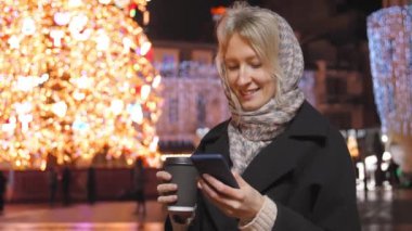 Genç ve çekici bir kadın akıllı telefon kullanıyor ve kışın dışarıda, Noel tatilinde, akşam vakti, Noel ağacının yanındaki meydanda kahve ikram ediyor. Parlak bulanık noel ışıkları.