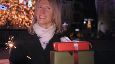 Güzel, stil sahibi, mutlu, pırıltılı bir kadın. Noel hediyeleriyle dolu bir kutu. Arka planda, kış gecesinde, tatil ışıkları ve Noel ağacı. Yeni yıl. İnsanlar kutlama yapıyor