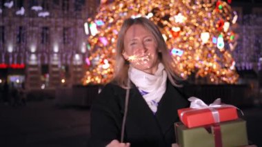 Güzel, stil sahibi, mutlu, pırıltılı bir kadın. Noel hediyeleriyle dolu bir kutu. Arka planda, kış gecesinde, tatil ışıkları ve Noel ağacı. Yeni yıl. İnsanlar kutlama yapıyor