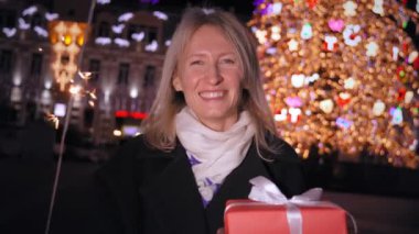 Güzel, genç, mutlu bir kadın elinde maytap ve hediyeler tutarak, sıcak ve neşeli bir atmosferde, şehir caddesinde, Noel ağacının yanında şenlikli bir kış akşamı neşe ve eğlence yayıyor. Ateş yanıyor..