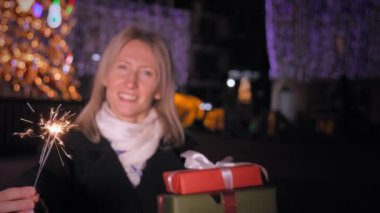 Güzel, genç, mutlu bir kadın elinde maytap ve hediyeler tutarak, sıcak ve neşeli bir atmosferde, şehir caddesinde, Noel ağacının yanında şenlikli bir kış akşamı neşe ve eğlence yayıyor. Ateş yanıyor..