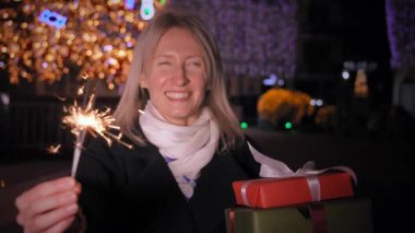 Güzel, stil sahibi, mutlu, pırıltılı bir kadın. Noel hediyeleriyle dolu bir kutu. Arka planda, kış gecesinde, tatil ışıkları ve Noel ağacı. Yeni yıl. İnsanlar kutlama yapıyor