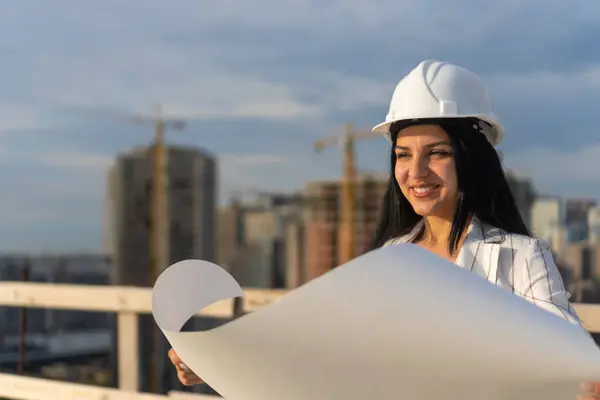 Gülümseyen genç bir mimarın portresi, tasarım mühendisi, bir inşaat sahasının üst katındaki hangi adamın kağıdına çizim yapılmış sert şapkalı bir müdür..