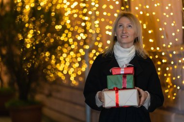Elinde üç kutu hediyeyle gülümseyen sarışın kadın Noel için şehrin sokak süslemelerinin arka planına bakmış..