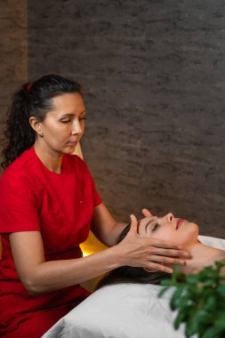 Kırmızı üniformalı bir masör spa solonunda masaj masasında yatan esmer bir kadına kafa masajı yapıyor. Spa konsepti, kişisel bakım. Dikey fotoğraf
