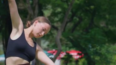 Genç bayan fitness eğitmeni parkta ısınıyor, yana eğiliyor ve çimlerin üzerinde diz çöküyor. Dışarıda antrenman yapmaya hazırlanıyor.