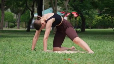 Spor giyimli genç bir kadın parkta yoga yapıyor, yoga minderinde kedi-inek pozu veriyor. Serin açık hava ayarları spor programını geliştirir.