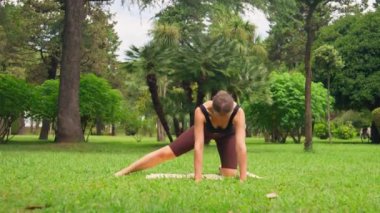 Spor giyim bölümünde genç bir kadın parkta yoga yapıyor. Paspasın üzerinde hareketli hareketler yapıyor. Serin açık hava ayarları spor ve rahatlamaya odaklanmasını sağlar.