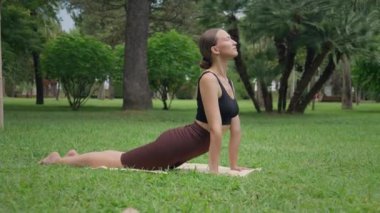 Spor kıyafetleri giyen genç bir kadın, yemyeşil parkta yoga yapıyor. Yoga minderinde farklı asmalar arasında geçiş yapıyor.