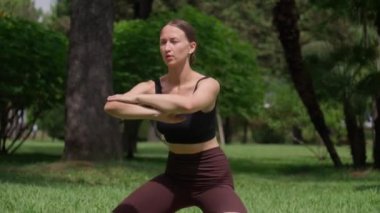 Yoga yapan genç bir kadın zengin parkında diz çökerken poz veriyor. Yeşilliğe gömülmüş, doğada zindelik ve sükuneti somutlaştırıyor. Nefes alma eğitimi..