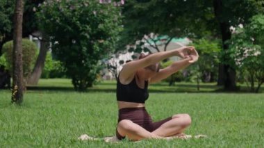 Spor giyimli genç bir kadın yemyeşil bir parkta yoga yapıyor. Esnekliğini ve farkındalığını vurgulayarak doğanın sükunetini kucaklıyor.