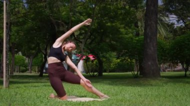 Spor hocası, yoga pozunun çeşitliliğini gösteriyor. Kollarını paspasın üzerinde sallıyor. Huzur parkı ortamında yemyeşil, sağlık ve refahı teşvik ediyor.