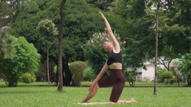 Spor hocası, yoga pozunun çeşitliliğini gösteriyor. Kollarını paspasın üzerinde sallıyor. Huzur parkı ortamında yemyeşil, sağlık ve refahı teşvik ediyor.