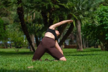Spor elbiseli genç bir kadın yeşil bir parkta yoga yapıyor. Kolunu arkaya doğru uzatarak doğa ile çevrili bir yoga minderinin üzerinde hareket ediyor. Akıllı spor anlayışı, yoga, pilates