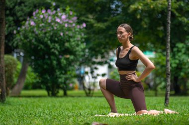 Beden eğitimi hocası, yemyeşil bir parkta diz çökmüş kalça esnekliği sergiliyor. Güneş ışığı ağaçların arasından süzülür, sağlık ve esneklik için sakin bir açık hava ortamı yaratır.