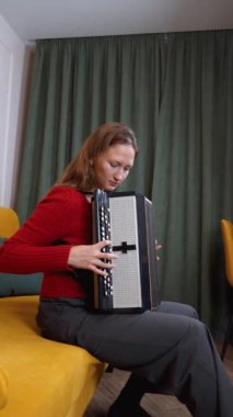 Kırmızı kazaklı kadın müzisyen sarı koltukta rahat bir şekilde otururken siyah beyaz akordeon çalıp sıcak ev ortamında melodik sesler çıkarıyor. dikey video