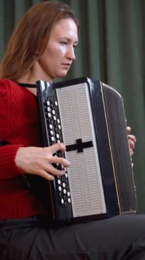 Kırmızı kazaklı genç bayan müzisyen son derece etkileyici taklit heyecanıyla akordeon çalıyor, profesyonel teknikle melodiyi seslendirirken müzik tutkusunu yakalıyor. dikey video