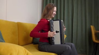 Kırmızı kazaklı kadın müzisyen sarı koltukta rahat bir şekilde otururken siyah beyaz akordeon çalıyor ve sıcak ev ortamında melodik sesler çıkarıyor..