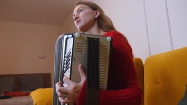 Kırmızı kazaklı kadın müzisyen sarı koltukta rahat bir şekilde otururken siyah beyaz akordeon çalıyor ve sıcak ev ortamında melodik sesler çıkarıyor..