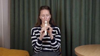 Kadın müzisyen, oturma odasındaki kanepede otururken flüt çalıyor ve doğal ışıklandırmalı küçük dekoratif masanın yanında rahat bir müzik performansı sergiliyor.