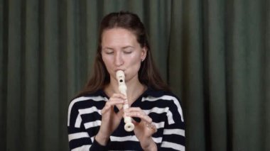 Kadın müzisyen, oturma odasındaki kanepede otururken flüt çalıyor ve doğal ışıklandırmalı küçük dekoratif masanın yanında rahat bir müzik performansı sergiliyor.