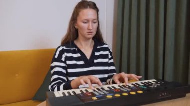 Kadın müzisyen elektronik klavyede melodik kompozisyon sergiliyor, rahat bir şekilde oturma odasındaki kanepede oturuyor, rahat, sanatsal bir ifadeyle.