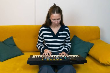 Genç bir kadın rahat sarı bir koltukta oturuyor, rahat ev ortamında müzik yaratmaya odaklanmış, elektronik bir klavye çalıyor.