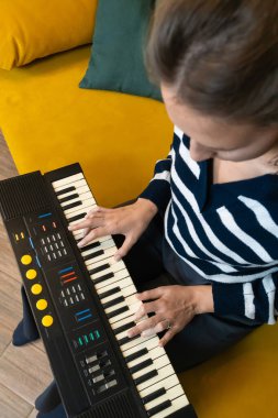 Kadın, canlı sarı bir koltukta rahat bir şekilde oturup müzik yaratma ve evde dinlenme anının keyfini çıkarırken tutkulu bir şekilde klasik bir sentezleyici çalıyor. Dikey fotoğraf