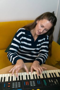 Kadın tutkuyla klasik bir sentezleyici çalıyor, arka planda sarı bir kanepe ile rahat bir ev ortamında müzik yaratıcılığı ve nostalji hissi uyandırıyor. Dikey fotoğraf