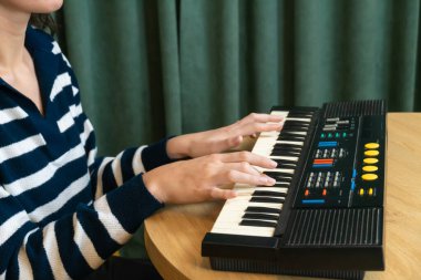 Genç bir kadın canlı sarı bir kanepede rahat bir şekilde oturuyor, modern dairesindeki ahşap bir masada klasik bir sentezleyici çalıyor, yaratıcılık ve sese dalmış bir şekilde.