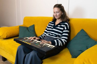 Modern bir oturma odasında rahat sarı bir koltukta oturan genç bir kadın, küçük bir elektronik klavye çalıyor, müzik yaratırken boş zamanlarının tadını çıkarıyor ve yaratıcılığını ifade ediyor.