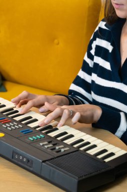 Sarı bir kanepenin yanında rahat bir şekilde oturan genç bir kadın, bir ev ortamında müzik yaratırken parmakları tuşların üzerinde dans ederek bir sentezleyici çalmaya kendini kaptırıyor. Dikey fotoğraf