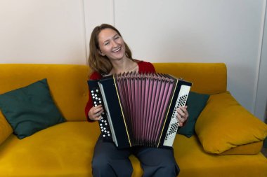 Kadın müzisyen gülümseyip geleneksel bir akordeon çalarken oturma odasındaki sarı kanepede rahatça oturup müzik anının tadını çıkarıyor.