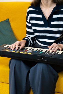 Kadın, canlı sarı bir koltukta rahat bir şekilde oturup müzik yaratma ve evde dinlenme anının keyfini çıkarırken tutkulu bir şekilde klasik bir sentezleyici çalıyor. Dikey fotoğraf