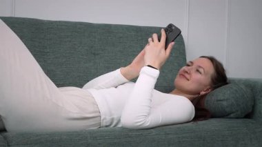 Yeşil kanepede rahat bir şekilde uzanan, beyaz uzun kollu gömlek giyen ve rahat ev ortamında dinlenirken akıllı telefonuyla meşgul olan genç bir kadın.