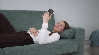 Genç bir kadın rahat bir kanepede dinleniyor, akıllı telefonuyla meşgul olurken evde huzurlu boş zamanların tadını çıkarıyor, modern dijital yaşam tarzını ve kişisel dinlenme zamanını temsil ediyor.