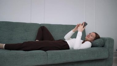 Genç bir kadın rahat bir kanepede dinleniyor, akıllı telefonuyla meşgul olurken evde huzurlu boş zamanların tadını çıkarıyor, modern dijital yaşam tarzını ve kişisel dinlenme zamanını temsil ediyor.